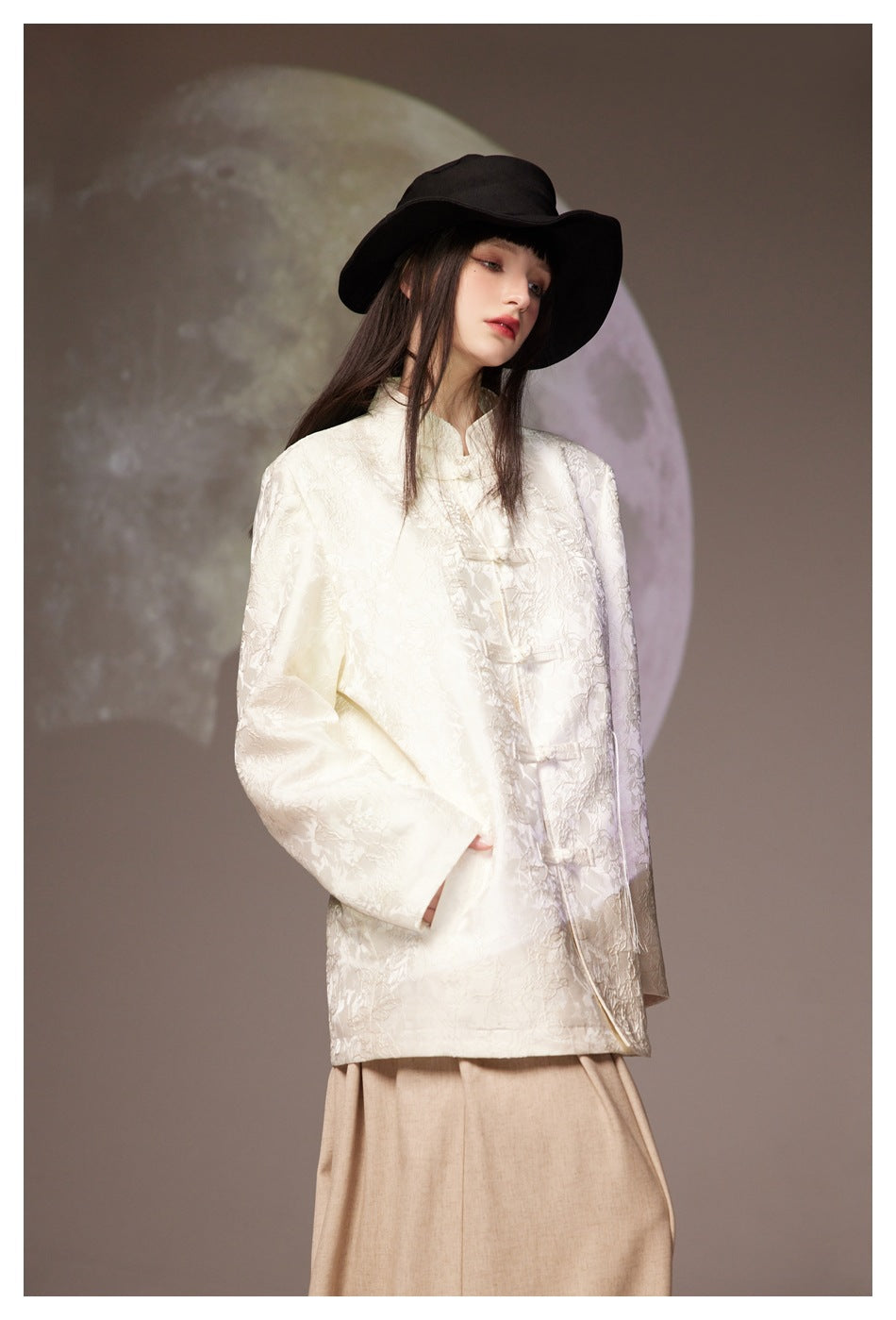 Angled view of a model wearing an ivory floral-textured jacket with frog closures and a black wide-brim hat.