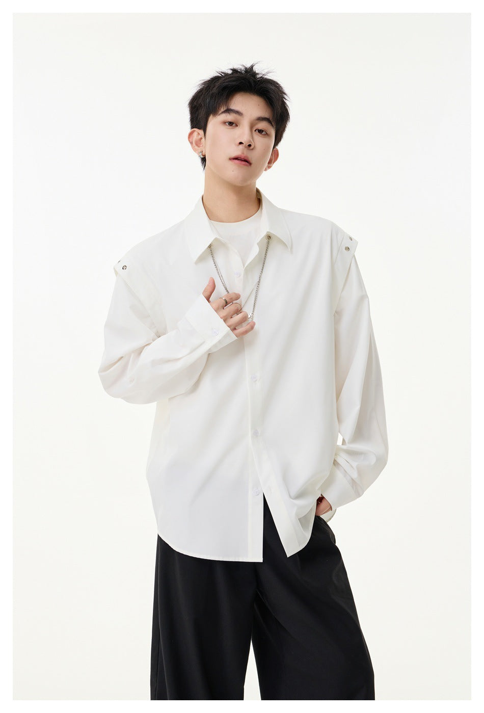 Model wearing an oversized white button-up shirt with snap-tab shoulders, layered chain necklace and black trousers.