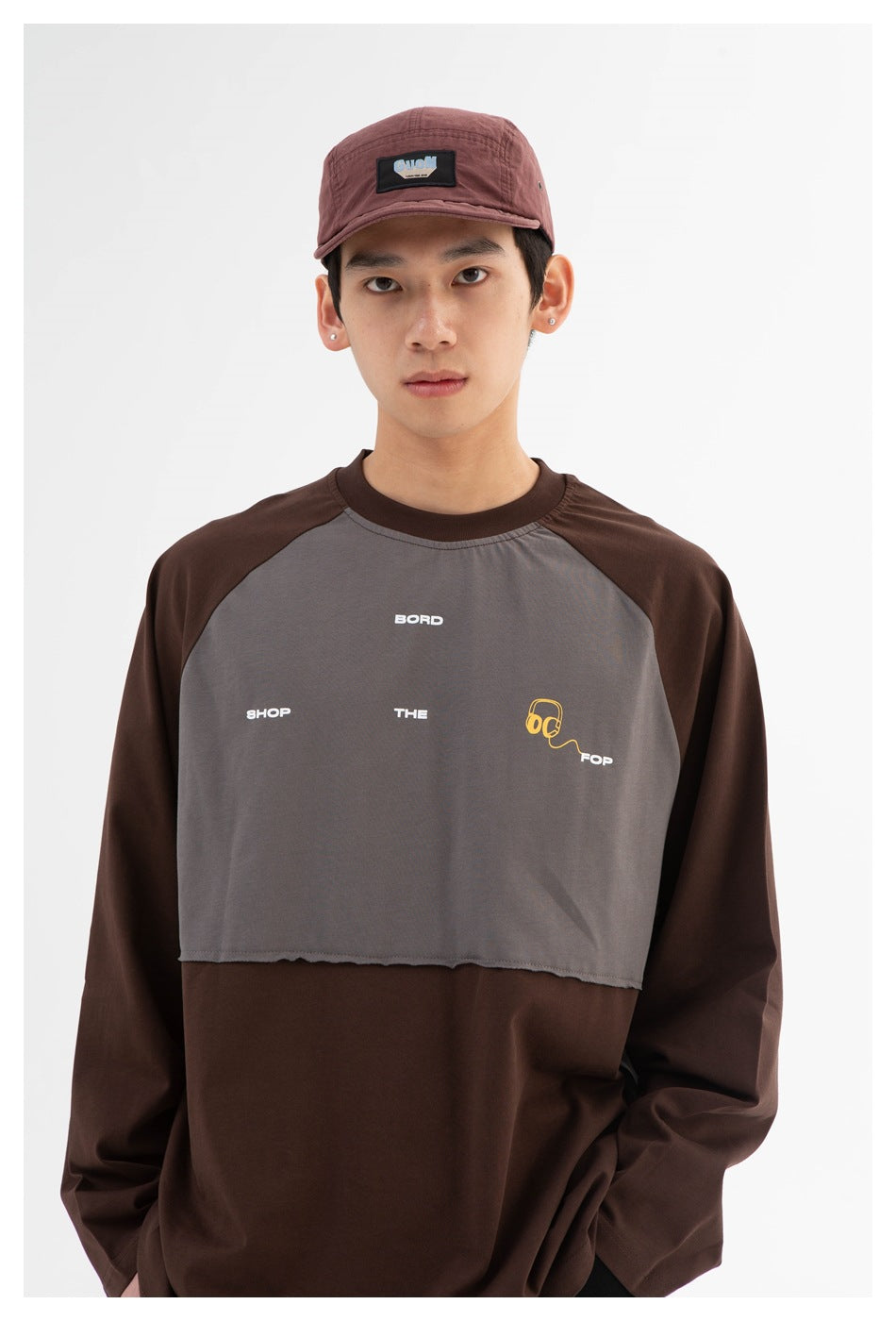 Front view of a model wearing a brown and gray color-block shirt with small chest graphics and a burgundy cap.