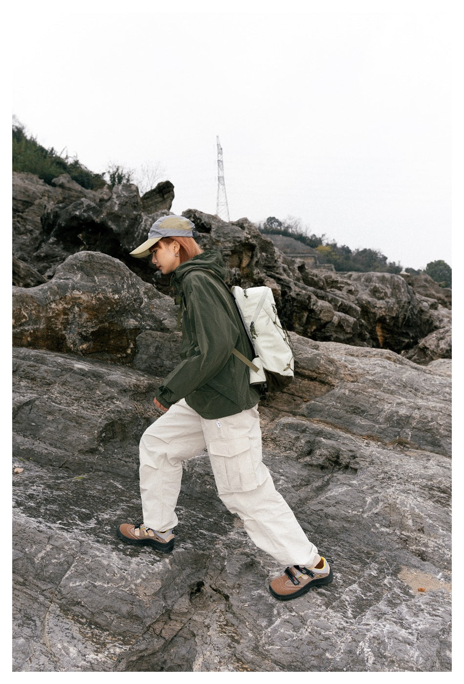 Model hiking over rocks in a dark green hooded jacket, cream cargo pants, and a gray-and-beige cap.