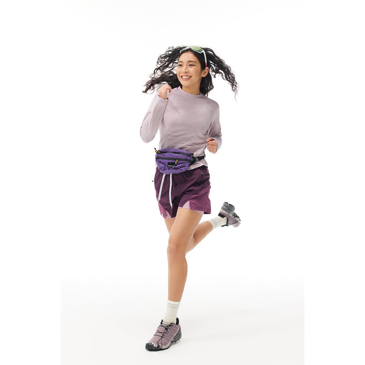 Model running in a pale mauve long-sleeve top, plum running shorts and a purple waist pack.