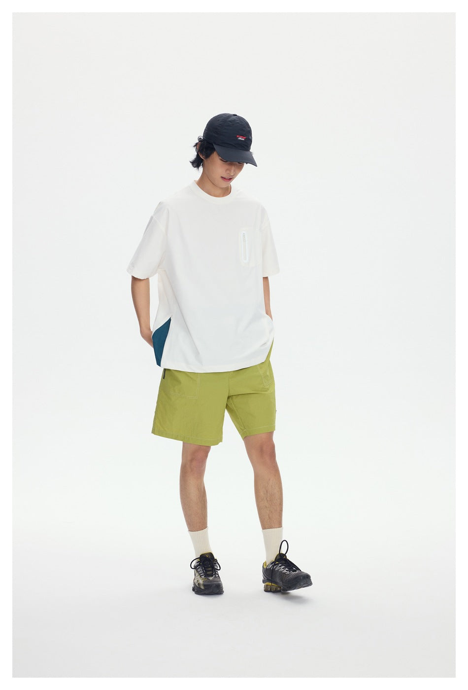 Model standing in green drawstring shorts with a white short-sleeve tee, black cap and sneakers