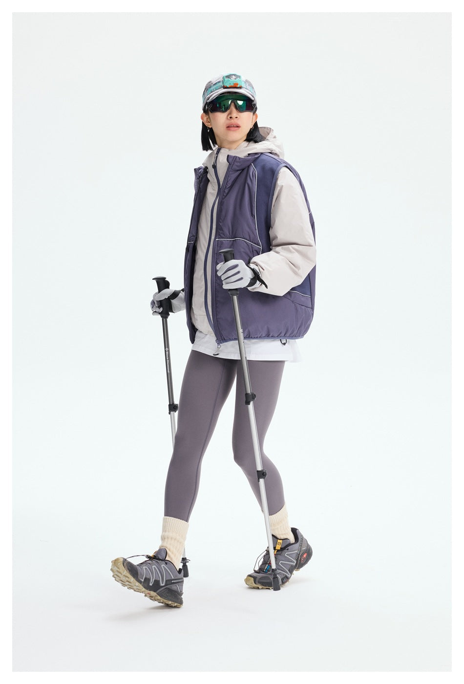 Model walking with trekking poles in a purple padded vest over a light hooded jacket and leggings