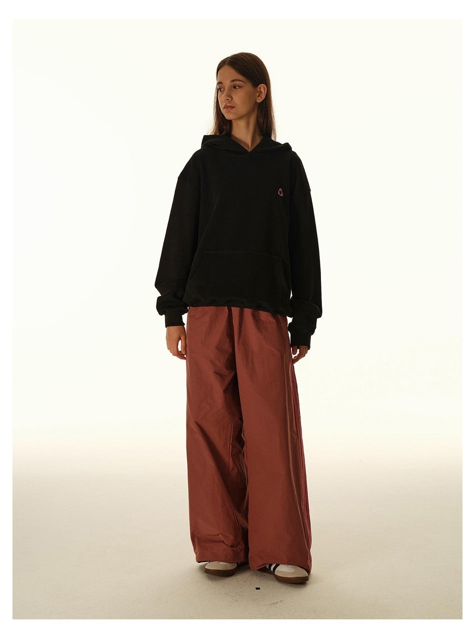 Model wearing a black hooded sweatshirt with a kangaroo pocket and small pink chest emblem, with rust wide-leg pants.