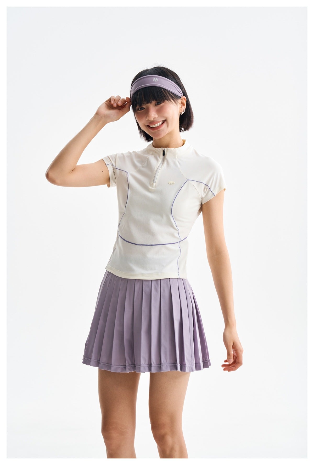 Model wearing a white short-sleeve quarter-zip top with lilac piping and a pleated lilac skirt.