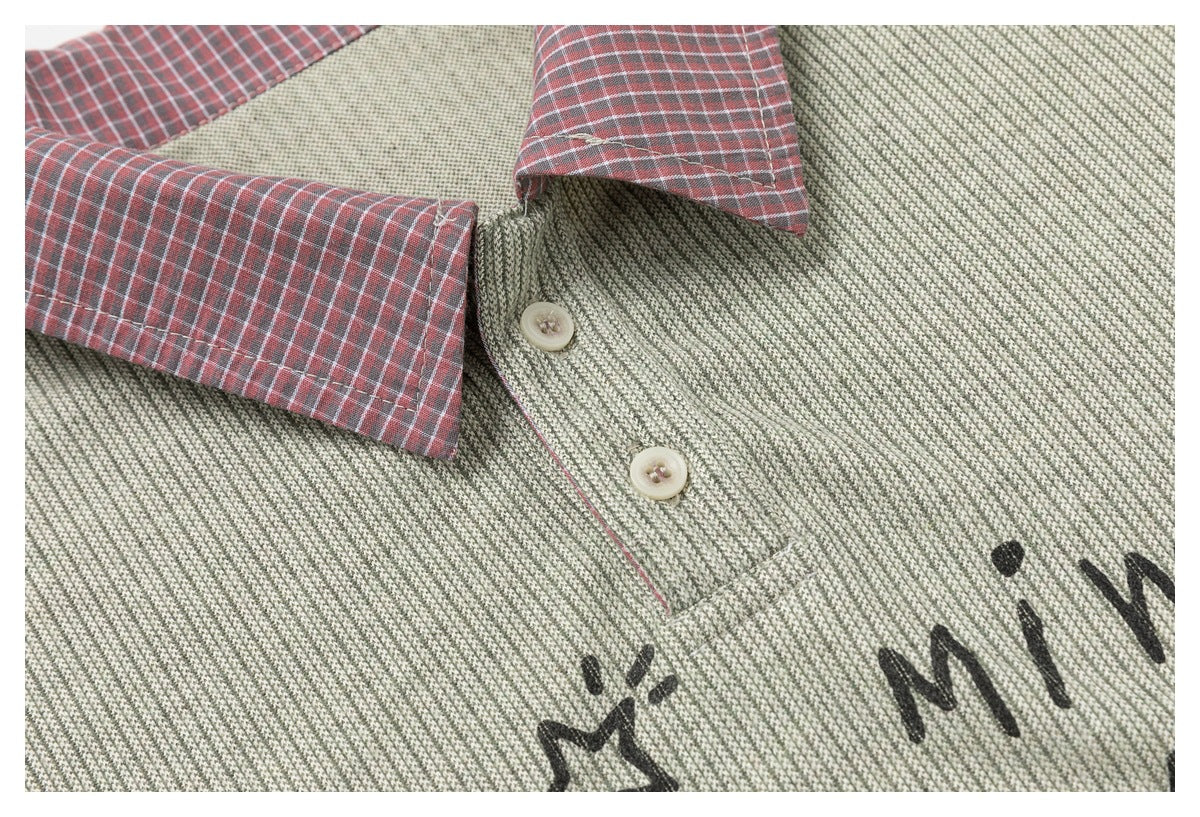 Close-up of the shirt's red plaid collar, cream buttons, sage-gray textured fabric, and partial black lettering.