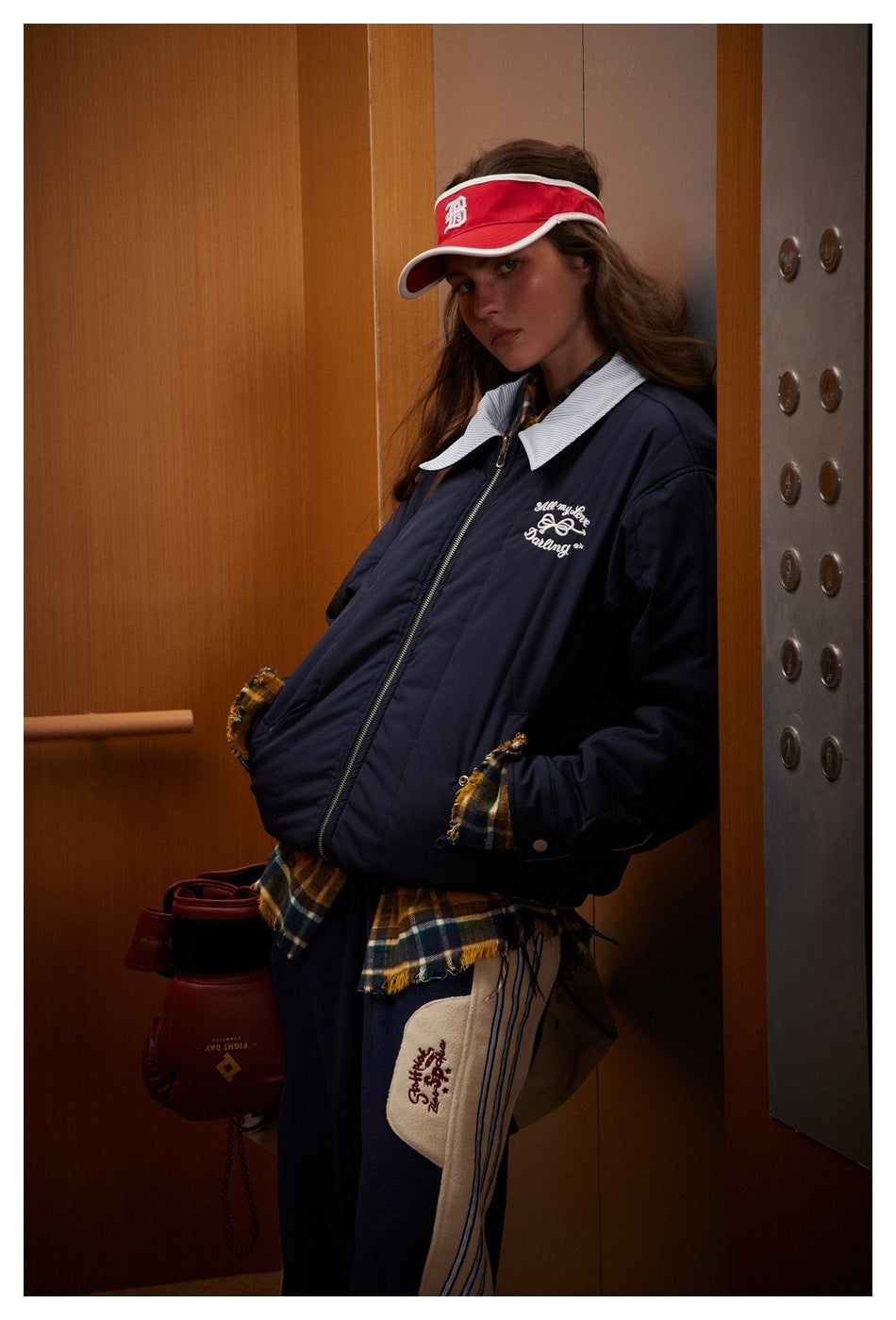 Model leaning in an elevator wearing the navy jacket with white script embroidery and striped collar, hands in pockets