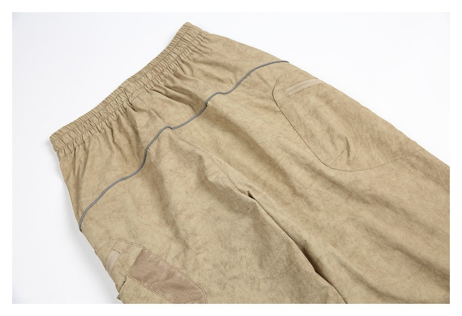 Partial flat lay of tan trousers with an elastic waistband, gray piping, and a mesh panel.