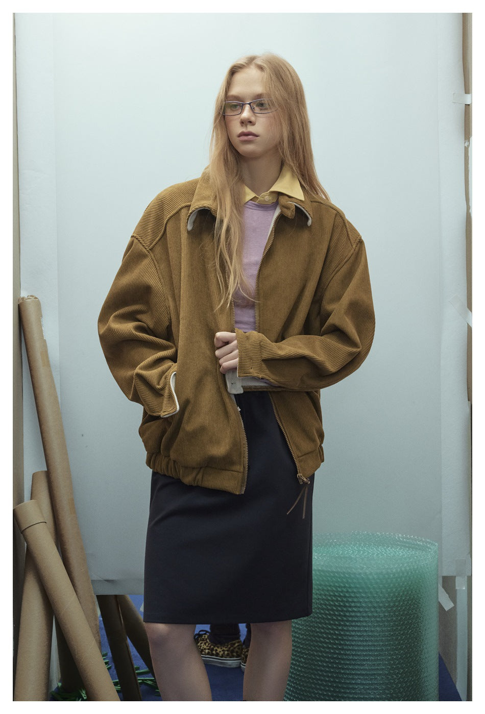 Model wearing an open brown ribbed zip jacket with a collar, hand at the front, over a lilac top and dark skirt