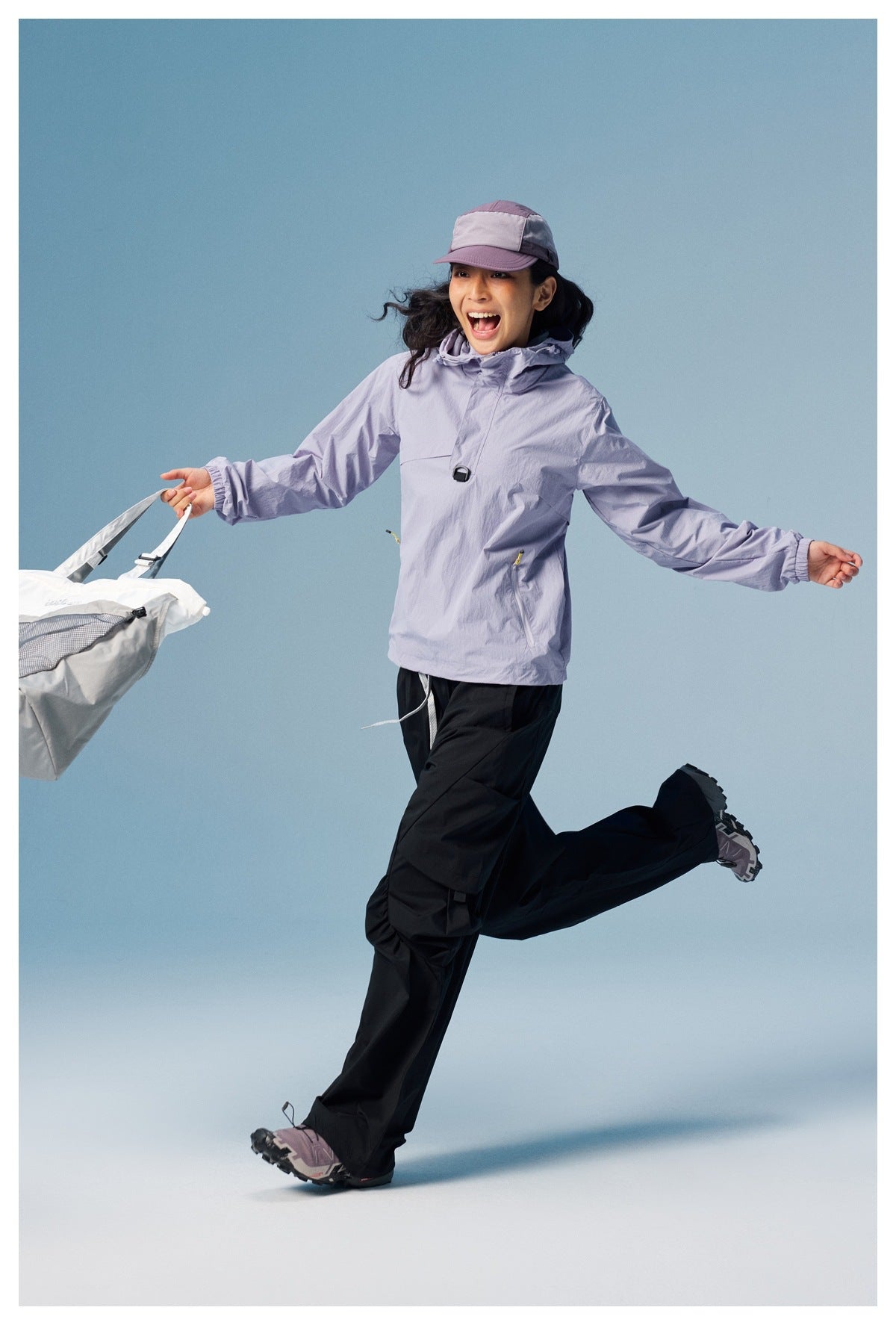 Model wearing a light lavender hooded pullover and black pants, with a matching cap, carrying a white bag.