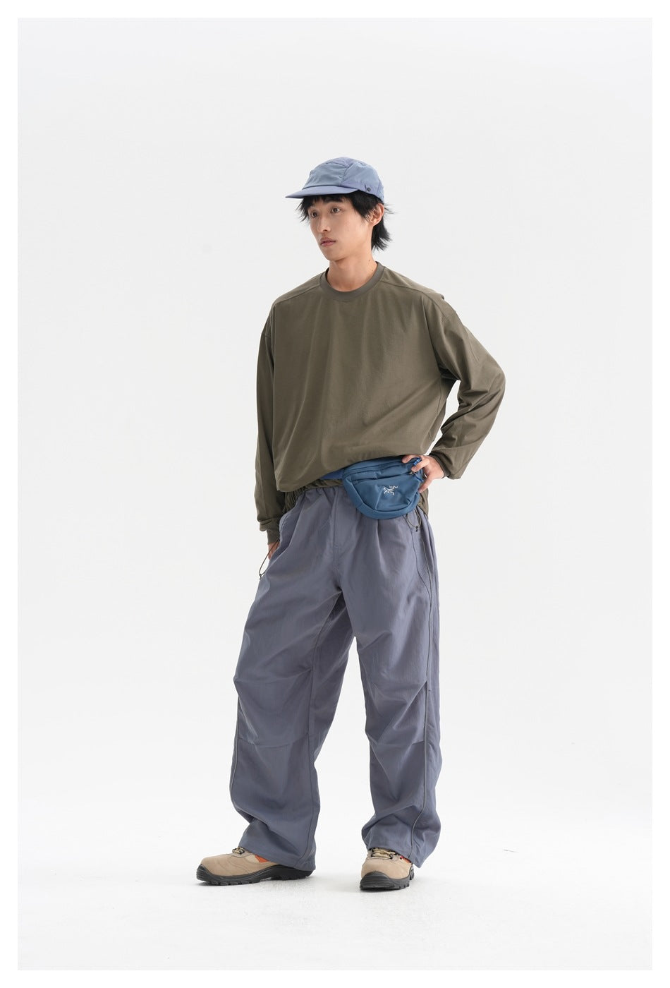 Man standing in an olive long-sleeve shirt and slate-blue wide-leg trousers, with a blue cap and waist bag.