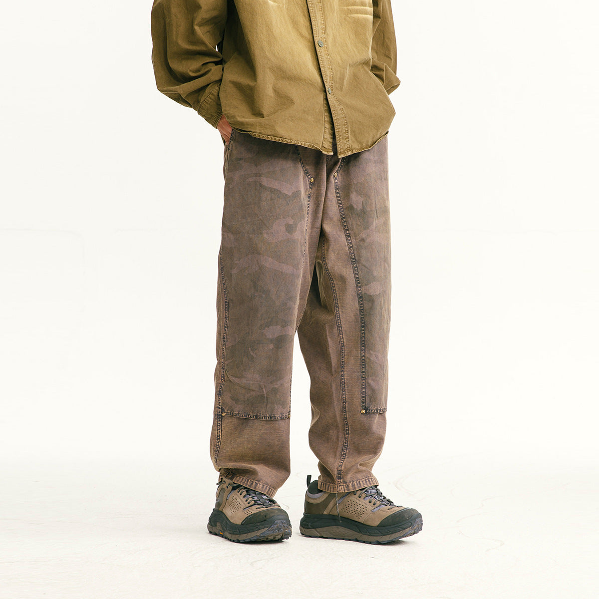 Cropped model wearing loose brown camouflage-panel pants with stitched seams and hiking shoes, shown from the front.