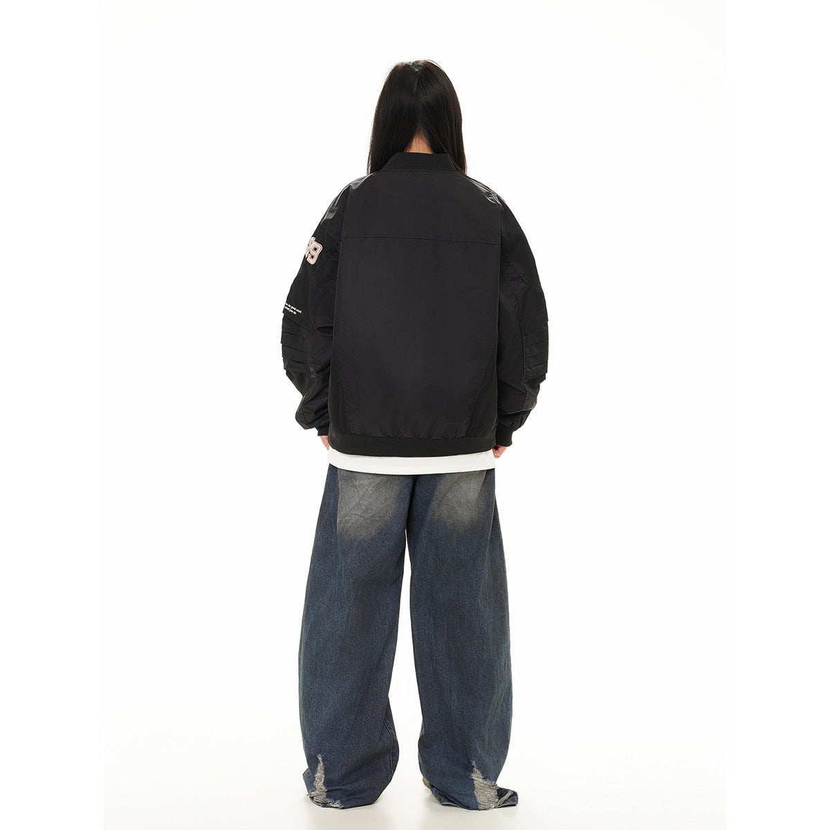 Rear view of a model wearing a black bomber jacket with sleeve patches and loose dark jeans with distressed hems.
