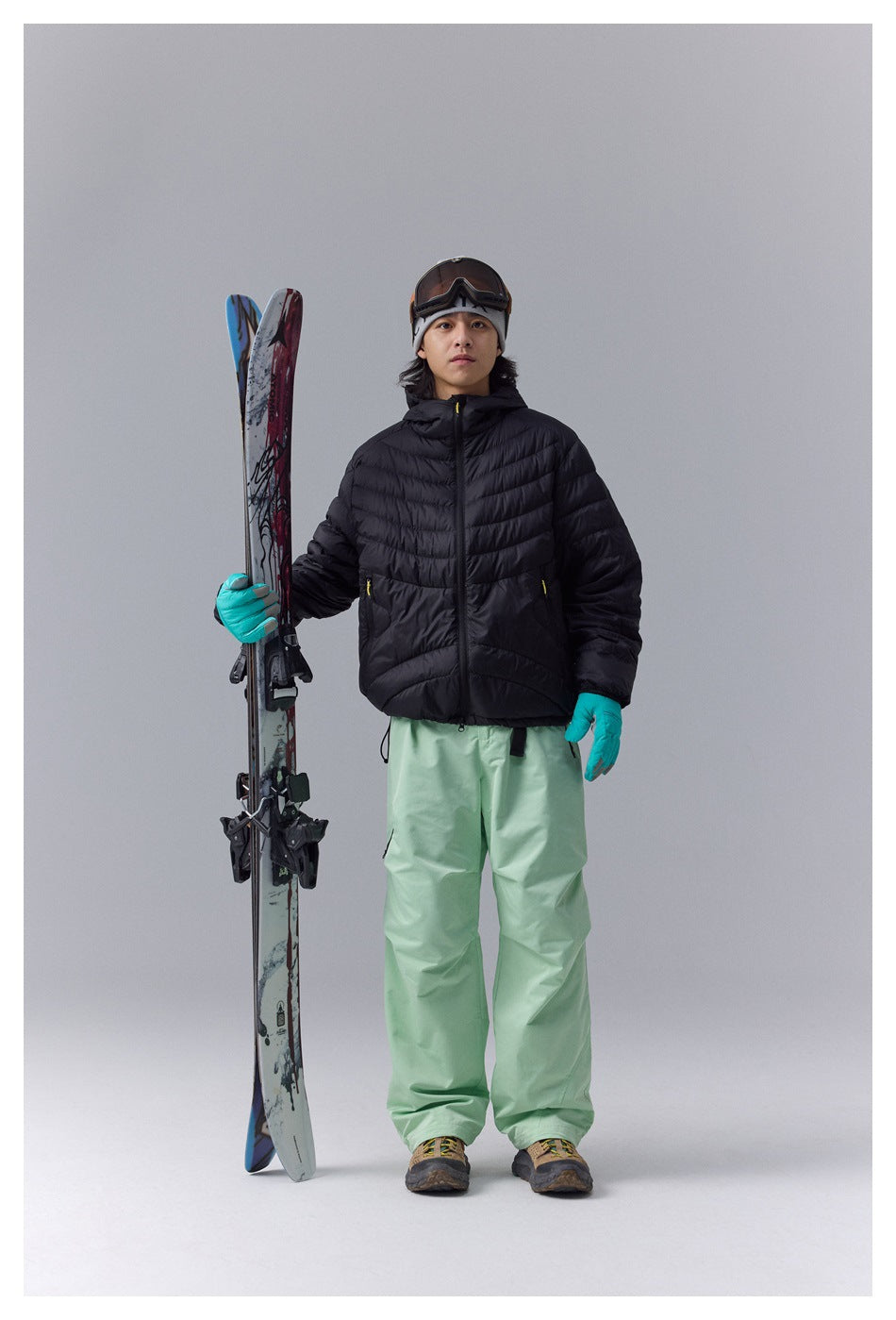 Model wearing a black quilted hooded jacket and pale green snow pants, holding skis.