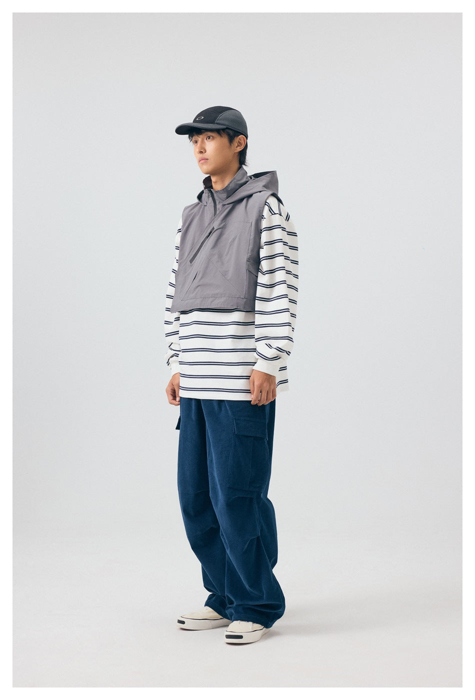 Model in gray hooded vest with diagonal zipper over a striped top and navy cargo pants, side view