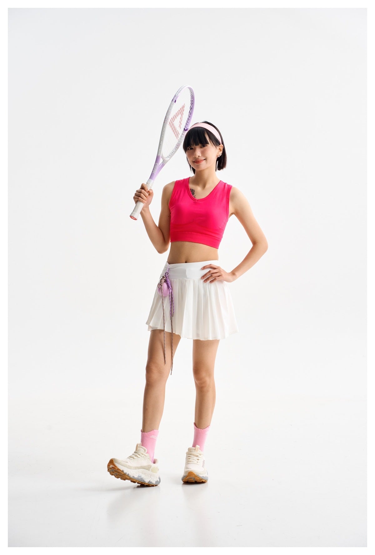 Model holding a tennis racket, wearing a bright pink crop top and white pleated tennis skirt.