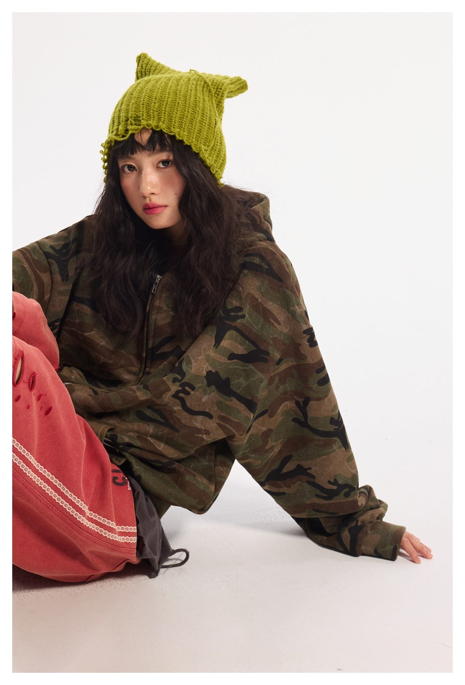 Model wearing an oversized brown, olive and black camouflage zip hoodie with a bright lime knitted pointed-ear hat.