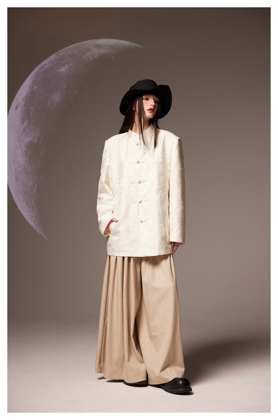 Model wearing an ivory frog-button jacket, beige wide-leg bottoms, and a black wide-brim hat against a moon backdrop.