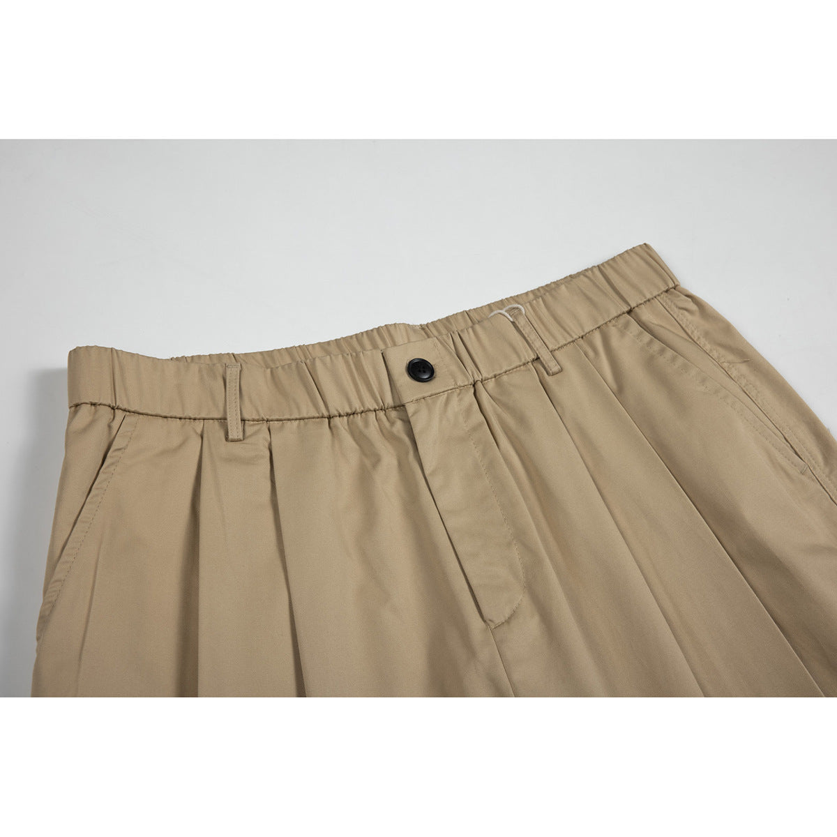 Close-up of tan trousers showing a gathered waistband, belt loops, black button, pleats and front pocket opening.