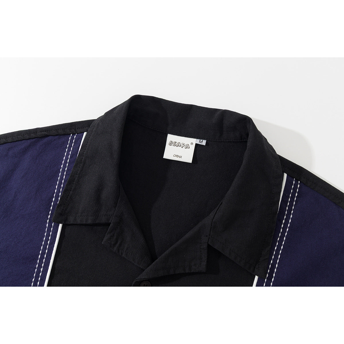 Close-up of a black collared shirt with navy shoulder panels, white piping and double rows of stitching; neck label visible.