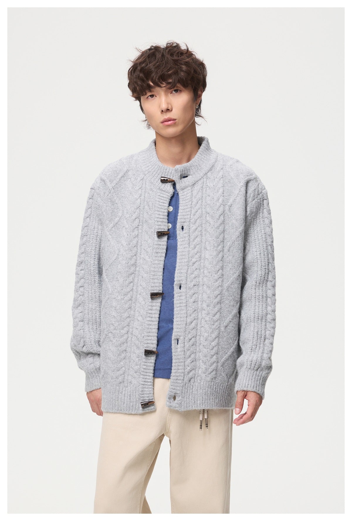 Front view of a model wearing a light gray cable-knit toggle cardigan over a blue shirt and cream trousers.