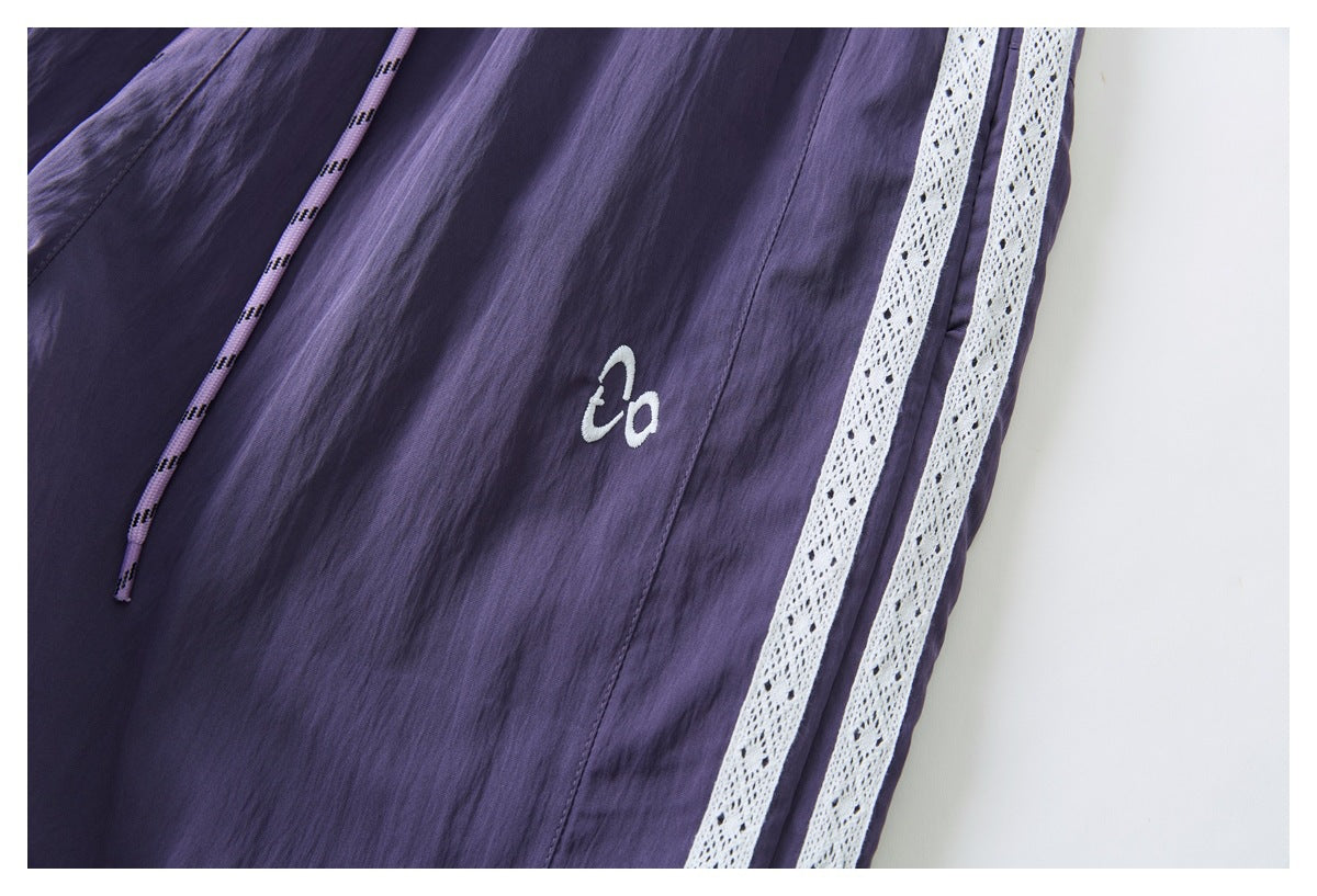 Close-up of purple pants with a white embroidered mark, patterned drawcord, and twin white lace side stripes.