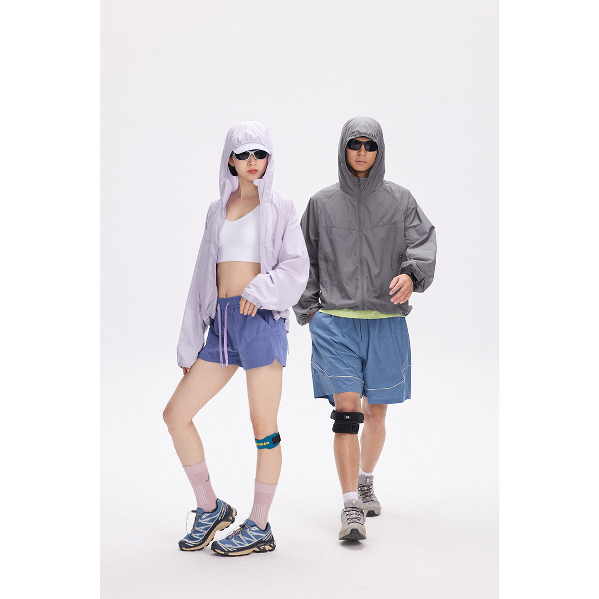 Two models wear hooded windbreakers and blue athletic shorts; one jacket is lavender and the other gray.