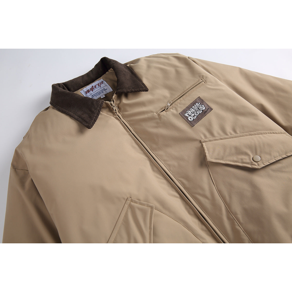 Close-up of a tan jacket front with a dark brown collar, zip closure, chest pocket and sewn patch.