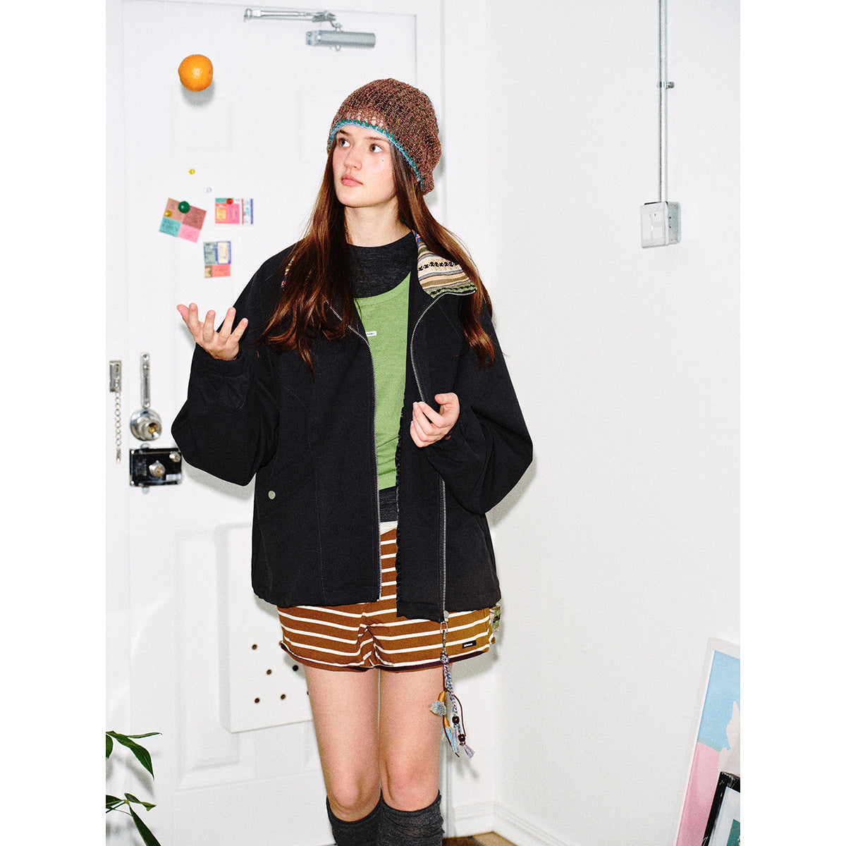 Model wearing an oversized black zip jacket with a patterned collar, striped shorts, and a brown multicolored knit beanie.