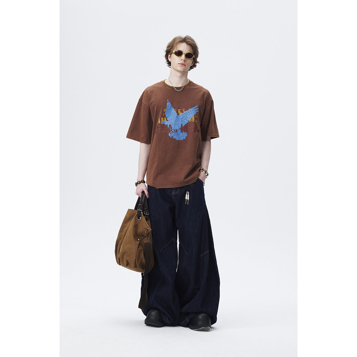 Model wearing an oversized brown T-shirt with a blue eagle graphic and very wide dark jeans.