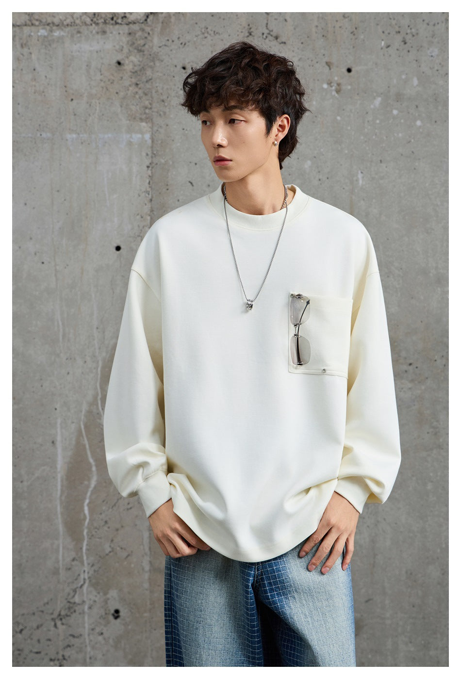 Model wearing an oversized ivory sweatshirt with a large chest pocket and relaxed sleeves; sunglasses are clipped to the