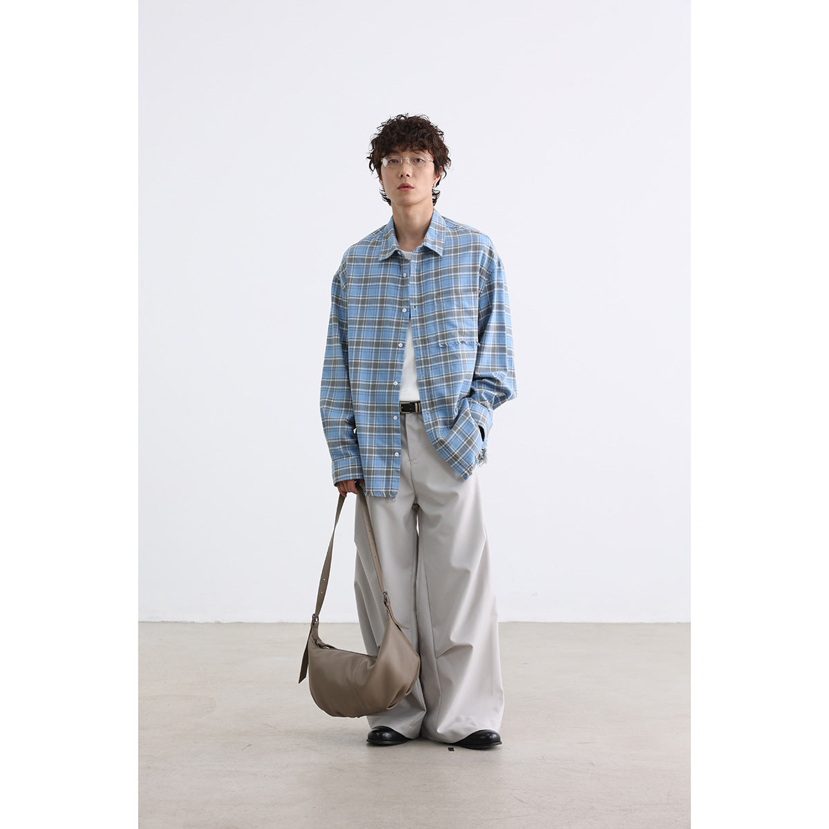 Model wearing a blue plaid button-up shirt with a chest pocket and loose light trousers, holding a shoulder bag.
