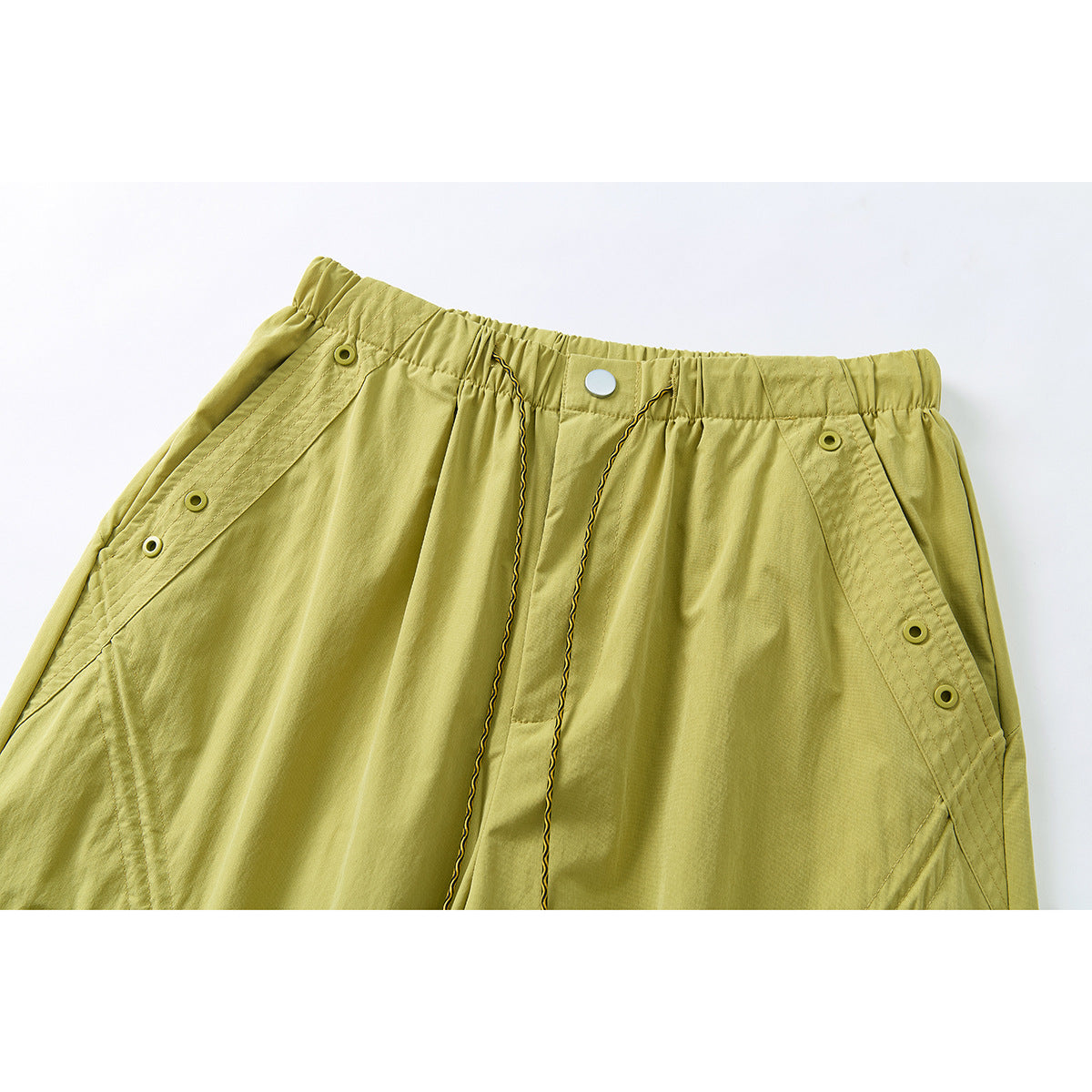 Close-up of the front waistband of mustard yellow-green trousers, showing a snap, drawcords and side button details.