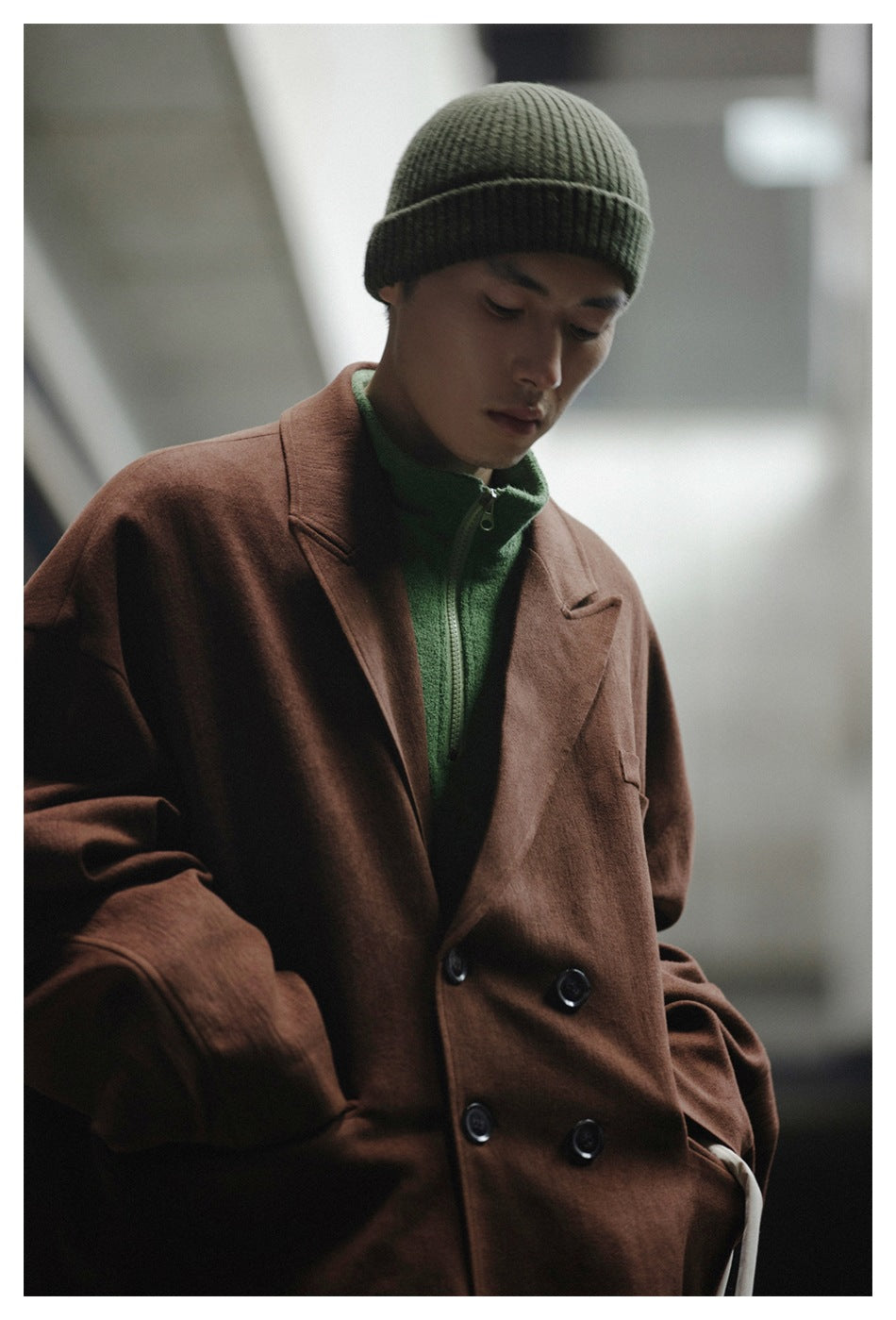Model wearing a brown double-breasted coat over a green zip-neck top and an olive ribbed beanie.