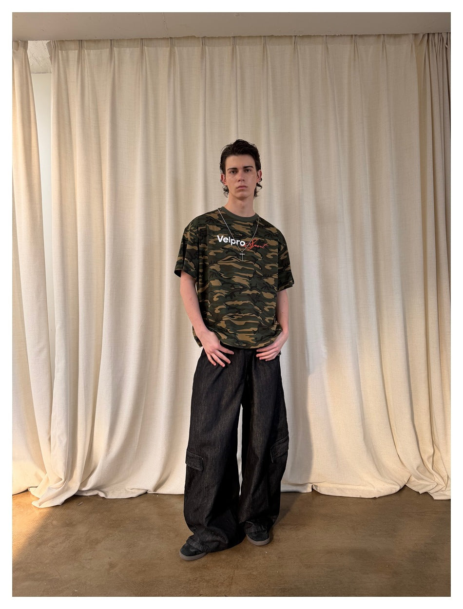 Full-length front view of a model wearing a green and brown camouflage graphic T-shirt with loose black cargo jeans.