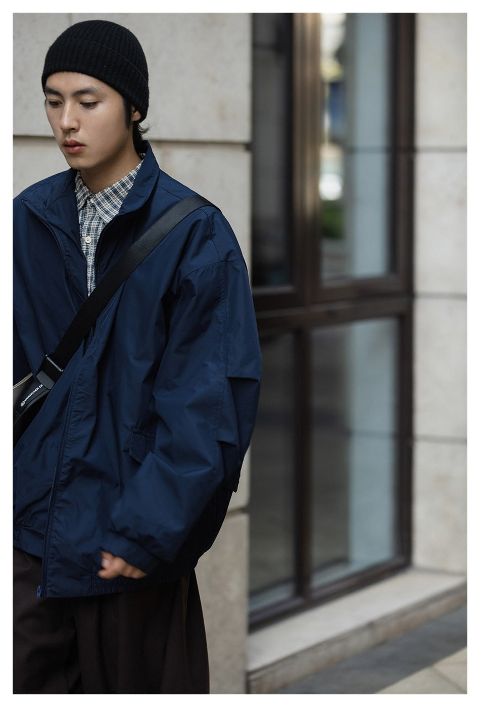 A man in a navy high-collar jacket and checked shirt wears a black knit beanie and crossbody bag.