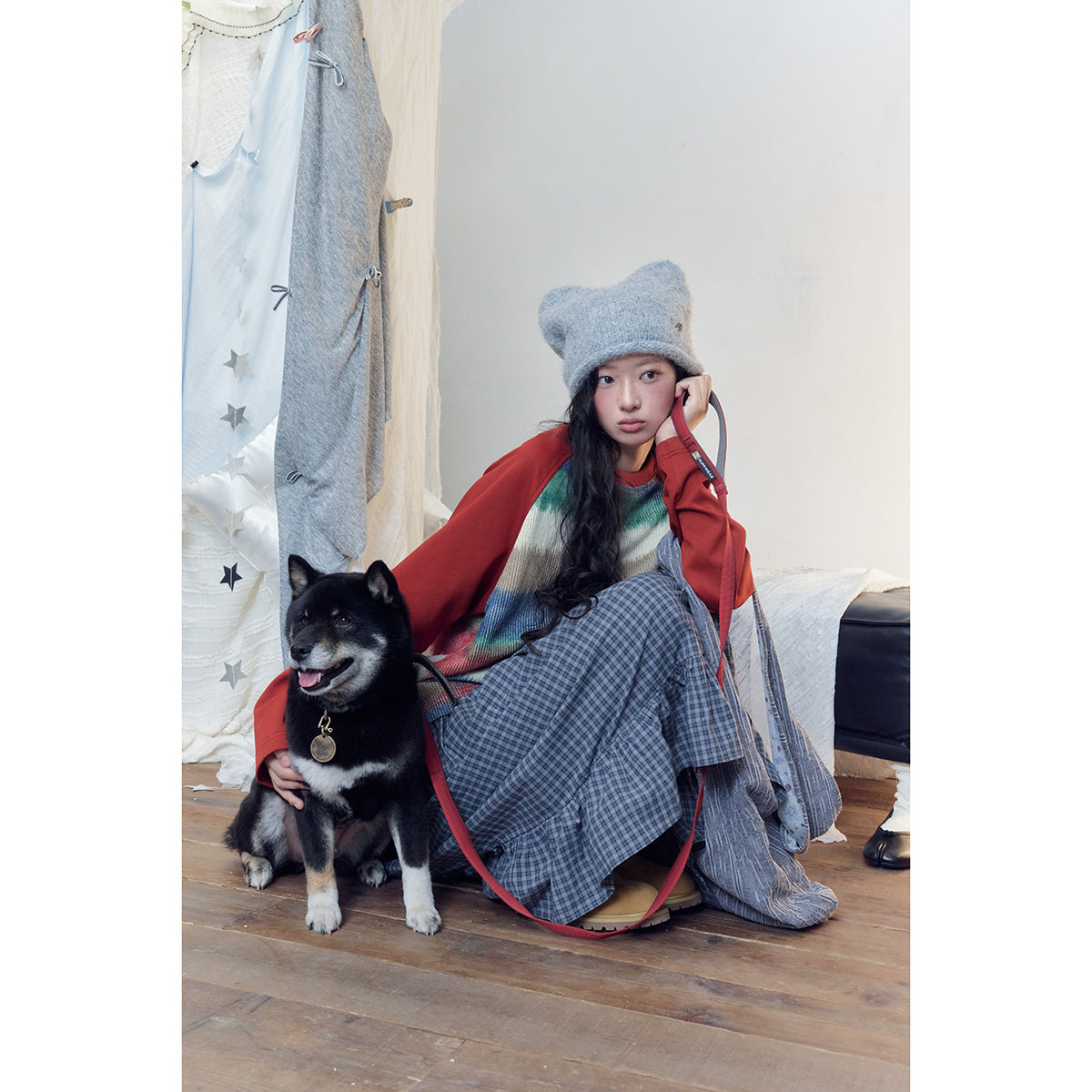 Model in a multicolored striped sweater, red outer layer, gray plaid skirt and fuzzy gray hat, seated beside a dog.
