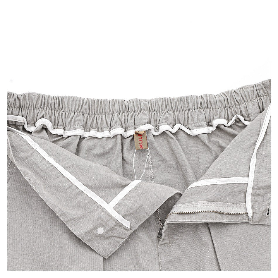 Close-up of gray trousers showing the gathered waistband, white ruffle trim, inner seams, and open zipper.