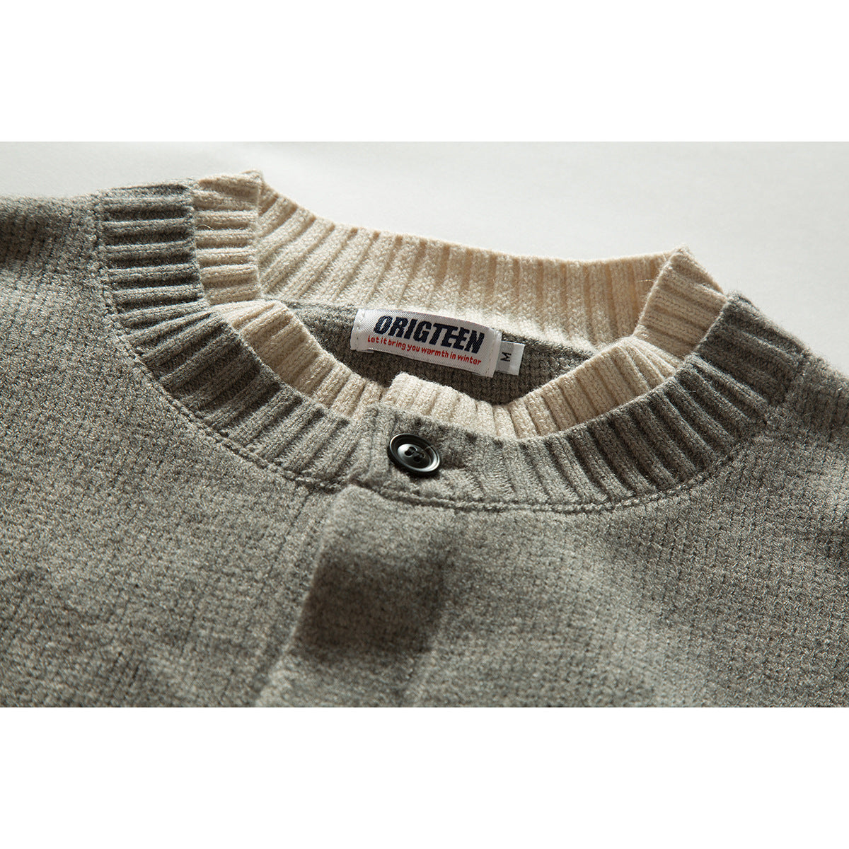 Close-up of a gray knit cardigan with a cream ribbed collar, black button, and sewn-in neck label.