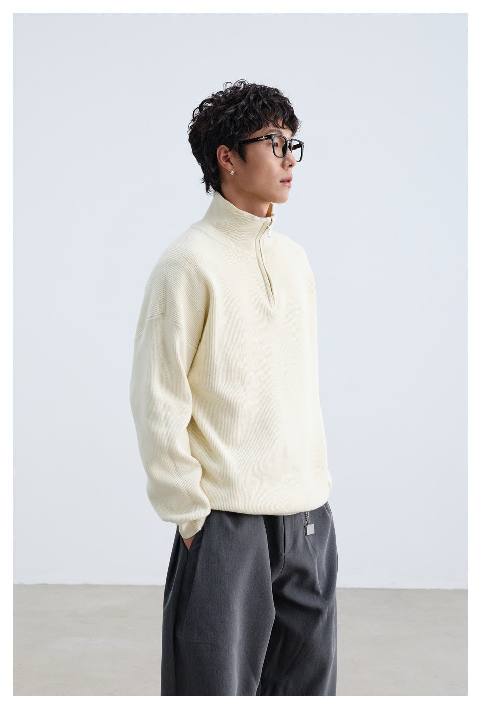 Model wearing an ivory quarter-zip pullover with a high collar and dark trousers, shown at a front angle.