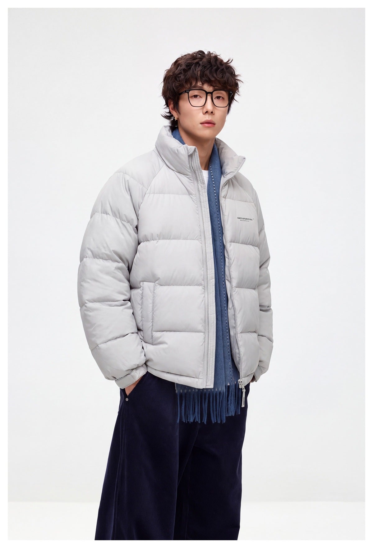 Model wearing a light gray quilted puffer jacket with a raised collar, blue scarf, and dark trousers.