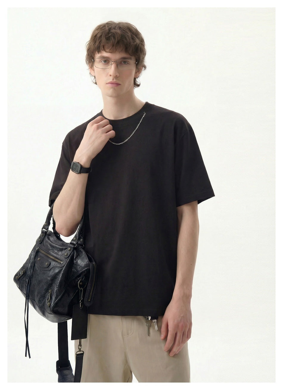 Man wearing a dark brown short-sleeve T-shirt with a neck chain, holding a black bag, with beige pants