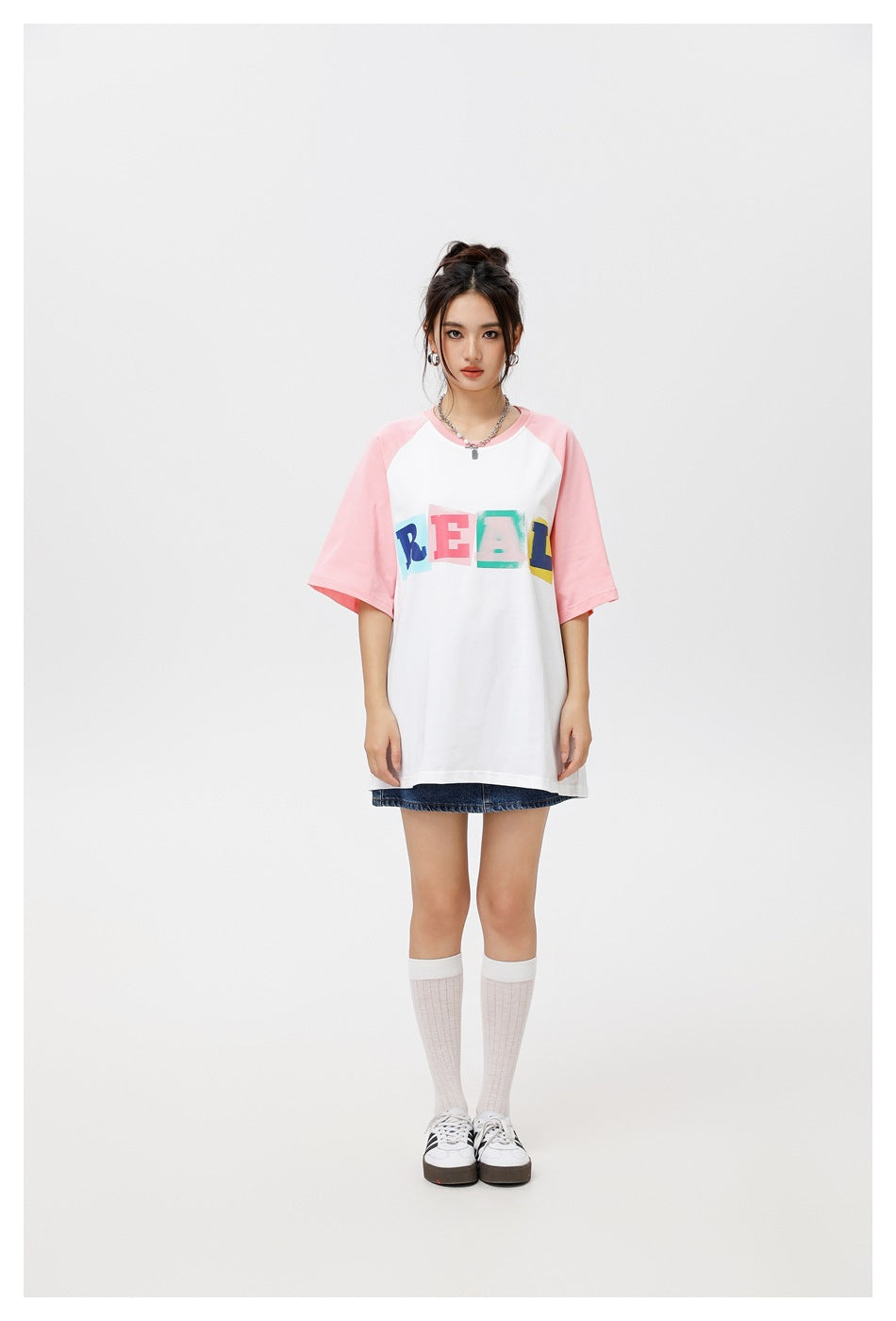 Model wearing a white oversized raglan T-shirt with pink sleeves and a colorful REAL print, with a denim skirt and white