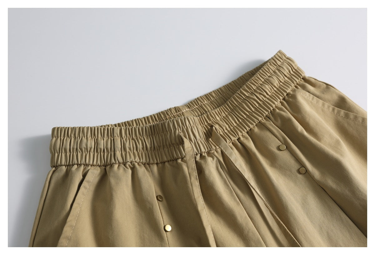 Close-up of tan shorts showing a gathered elastic waistband, drawstrings, pleats and small metal buttons.