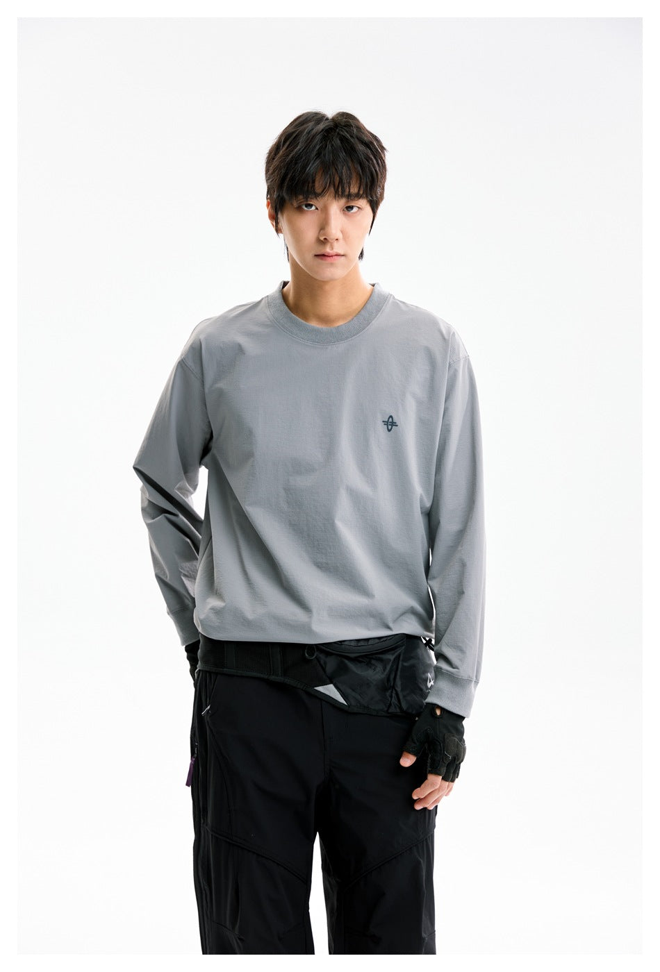Model wearing a gray long-sleeve crewneck with a small chest emblem and black pants, shown from the front.