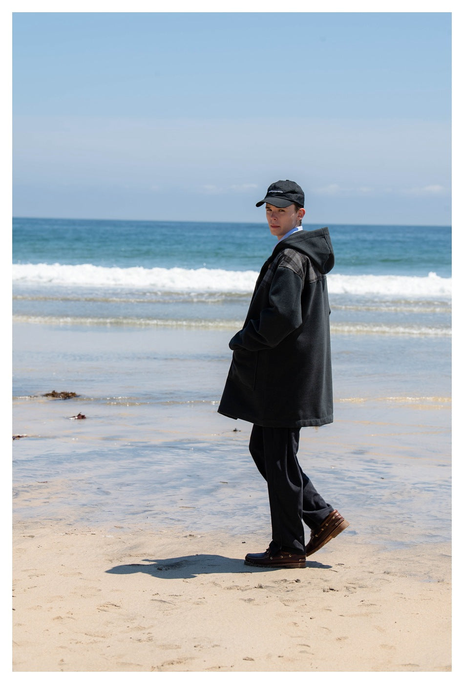 Model wearing a long charcoal hooded coat with a plaid shoulder panel, standing on a beach in side view.