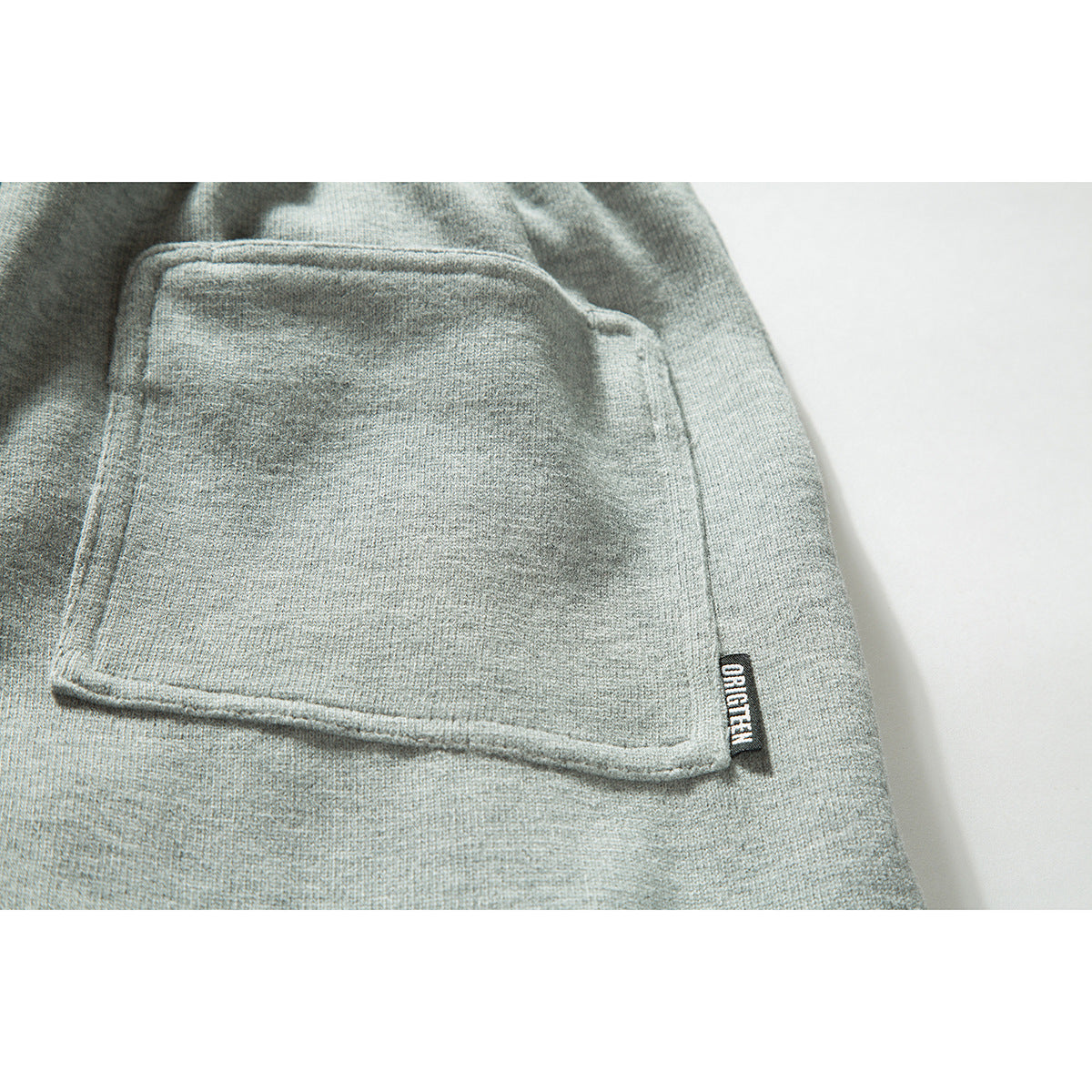 Close-up of a square pocket on light gray sweatpants with a small black sewn-in tag.