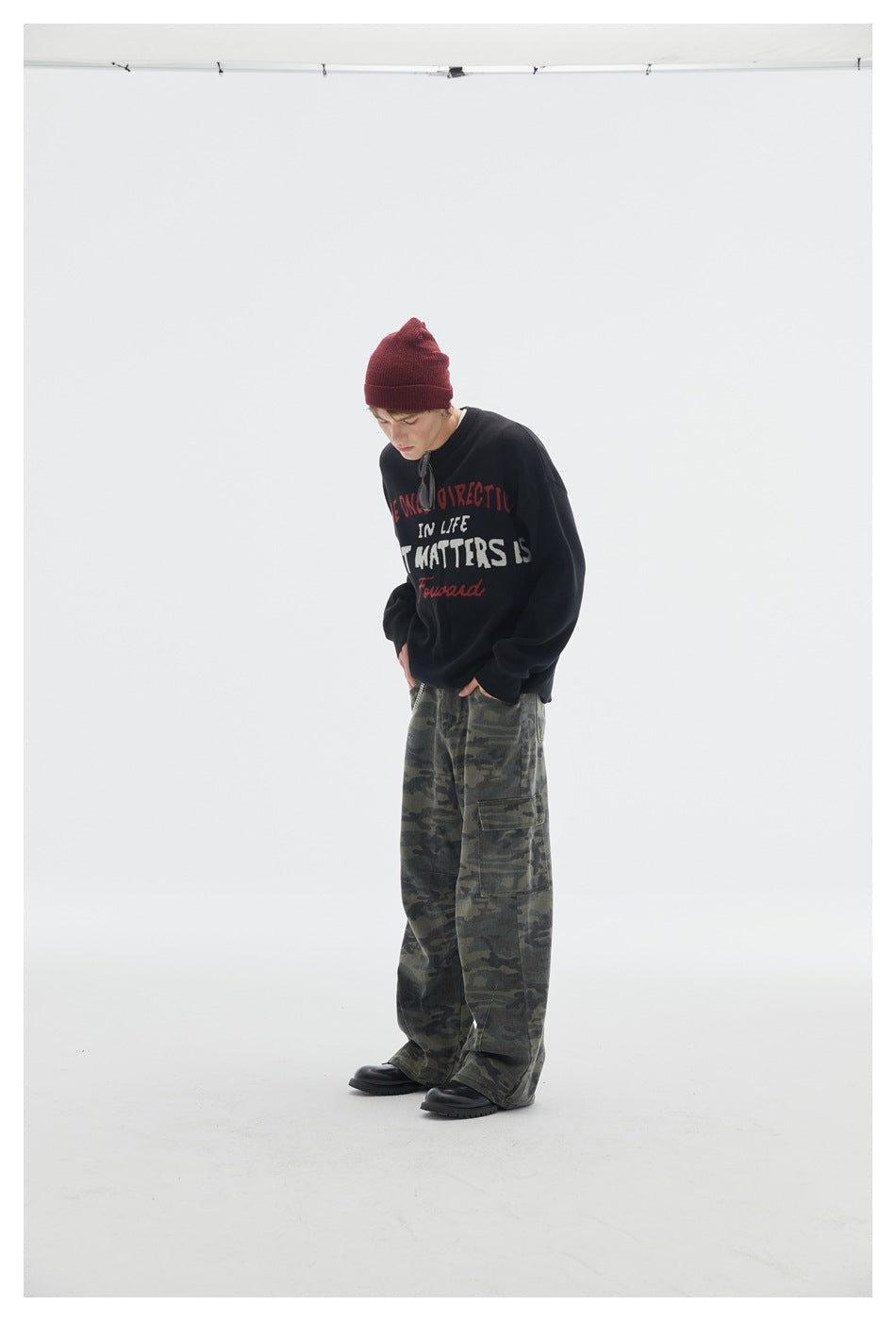 Model leaning forward in a black lettered crewneck sweater, burgundy beanie, and loose camouflage cargo pants.