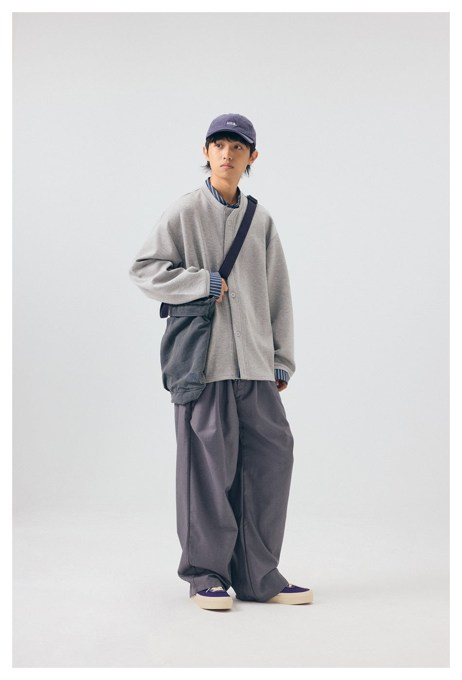 Person wearing the gray layered button-front jacket with wide gray trousers, cap and shoulder bag, front view
