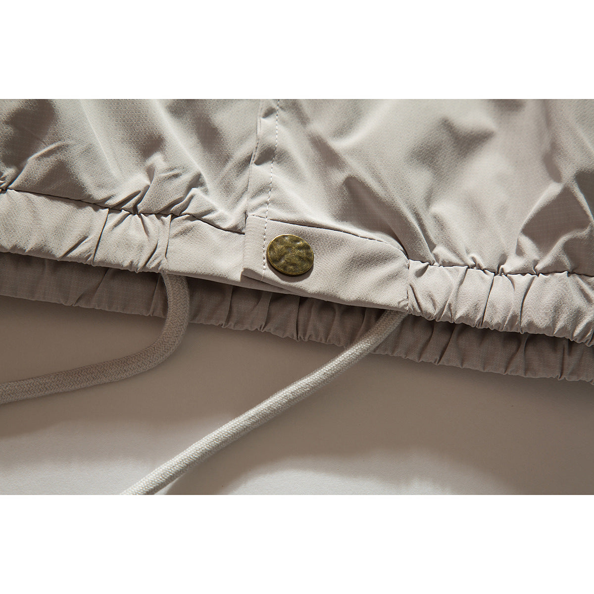 Close-up of a light beige jacket's gathered hem, drawcord, and brass-tone button tab.